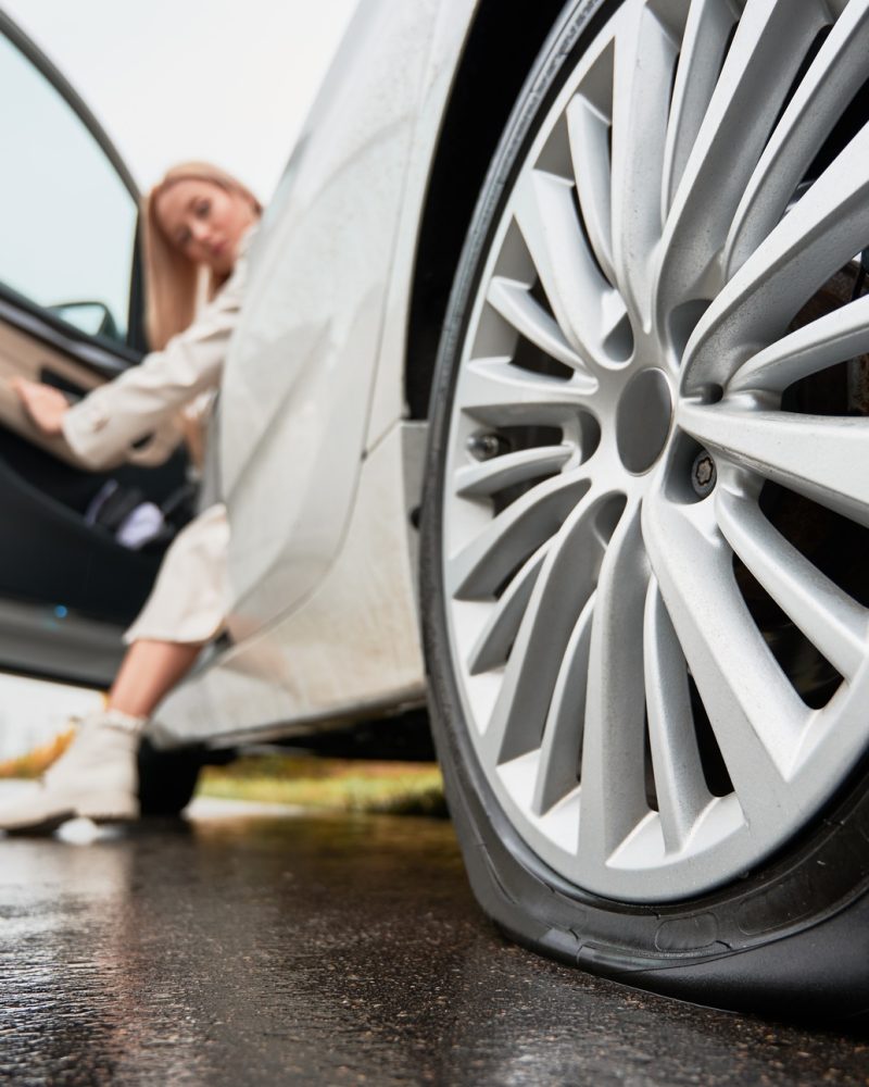 Female driver of white car observing punctured tire on rear wheel.